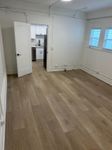 Bright and modern room with elegant wood flooring, large windows, and a minimalistic kitchen visible through an open door.