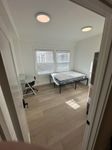 Bright bedroom featuring a comfortable bed, modern desk setup, and natural light streaming through large windows.