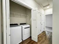 Modern in-unit laundry area with sleek washer and dryer, integrated shelving, and stylish wooden flooring leading to a cozy carpeted room.