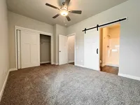 Spacious bedroom featuring plush carpeting, a sleek ceiling fan, and a modern barn door leading to an elegant en-suite bathroom.