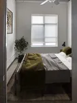 Cozy bedroom with modern decor, featuring a large window with blinds, stylish bed with green bedding, and a vibrant plant adding a touch of nature.