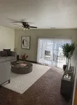 Bright and inviting living room with modern decor, plush carpeting, a stylish ceiling fan, and large sliding glass doors leading to a sunny outdoor space.
