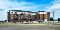 Modern brick apartment building with balconies, surrounded by a well-maintained parking area and landscaped greenery under a vibrant blue sky.