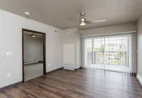 Bright and airy apartment living room with modern ceiling fan, large windows, and wooden flooring, leading into a cozy bedroom.