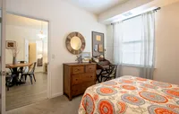 Spacious bedroom in a modern apartment featuring luxury carpeting, ample natural light, and a sleek, contemporary design for a comfortable and stylish retreat. Cozy bedroom with elegant decor, featuring a stylish wooden dresser, a work desk, and a vibrant patterned bedspread. Sunlight streams through sheer curtains, adding warmth to the inviting space.
