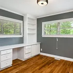 Bright home office with modern built-in desk, ample shelving, and large windows with lush green views.