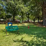 Charming outdoor play area set in a lush, sunlit park with vibrant playground equipment and towering trees.