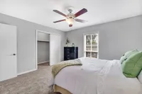 Bright and welcoming bedroom with soft neutral walls, featuring a cozy bed adorned with green pillows, a modern ceiling fan, and ample natural light streaming through a large window.