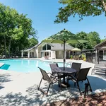 Inviting apartment complex with a sparkling pool, lush greenery, and cozy outdoor seating under a sun umbrella.