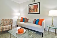 Inviting living room with a modern beige couch, vibrant orange and blue pillows, stylish patterned chair, and elegant glass tables, complemented by warm lighting and abstract wall art.