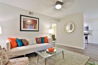 Elegant living room featuring a chic white sofa with vibrant, colorful pillows, a glass coffee table with decorative vase, and stylish artwork. Modern open-concept design with a glimpse into a sleek kitchen.