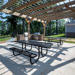 Charming outdoor patio with pergola, featuring picnic tables and a barbecue grill, set against a lush green backdrop.