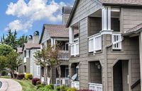 Charming residential apartment complex with quaint wooden balconies, lush greenery, and a serene pathway under a bright blue sky.
