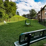 Inviting fenced dog park in apartment community with lush green grass, play tunnel, and decorative bench under a vibrant blue sky.
