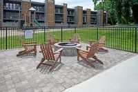 Charming outdoor patio with cozy Adirondack chairs surrounding a fire pit, set against modern apartment buildings and a lush green lawn.