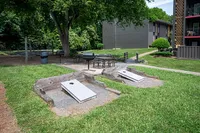 Charming outdoor area featuring a cornhole setup, picnic tables, and BBQ grills, surrounded by lush greenery and modern apartment buildings.