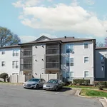 Modern apartment complex with three-story structure, multiple balconies, ample parking, and scenic greenery under a bright blue sky.
