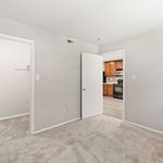 Bright and inviting apartment room with plush carpeting, leading to a modern kitchen featuring wooden cabinets and sleek appliances.