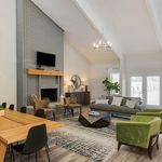 Elegant living room with high ceilings, modern chandelier, and cozy seating area featuring a stylish gray sofa and vibrant green chairs. The space includes a classic brick fireplace, large windows, and natural light creating a warm, inviting ambiance.