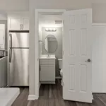Modern apartment view featuring a sleek stainless steel refrigerator next to a compact, stylish bathroom with elegant lighting and an oval mirror.
