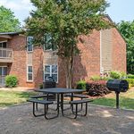 Charming apartment exterior with a cozy brick facade, complemented by a lush tree and a convenient outdoor picnic area with a barbecue grill.