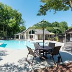 Inviting apartment complex with a sparkling pool, lush greenery, and cozy outdoor seating under a sun umbrella.