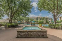 Luxurious apartment complex with a sparkling pool surrounded by lush greenery and elegant stone landscaping under a bright blue sky.