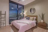 Modern bedroom with stylish decor, featuring a cozy bed with pink bedding, large windows showcasing a vibrant city view, and a sleek bedside table with elegant accessories.