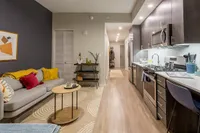 Modern apartment interior showcasing a cozy living area with a stylish gray sofa and vibrant cushions, seamlessly connected to a sleek, fully-equipped kitchen with contemporary cabinetry and stainless-steel appliances.