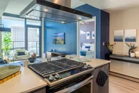 Contemporary apartment kitchen with sleek island stove, vibrant blue accent wall, and a cozy living area with large windows offering stunning city views.