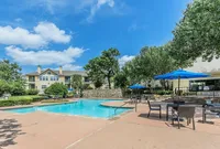 Inviting apartment complex with a sparkling pool, vibrant blue parasols, and lush greenery under a clear blue sky.