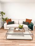 Style Meets Comfort Chic living room with a modern beige sofa adorned with colorful cushions, a stylish glass-topped coffee table, and vibrant potted plant accents on warm wooden floors.