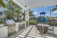 Grill with a view Modern outdoor kitchen and dining area with stainless steel grills and seating under a stylish pergola, next to a vibrant playground surrounded by lush greenery.