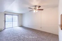 Bright and inviting living room in The Hampton apartment, featuring plush carpeting and a ceiling fan, ideal for comfort. Large window with vertical blinds offers abundant natural light in this spacious 1 bed, 1 bath layout.