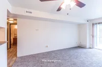 Bright and welcoming living space in The Hampton, featuring modern ceiling fan, plush carpet, and ample natural light from large windows. 1 bed, 1 bath, 630 sq. ft.