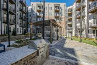 Modern apartment complex courtyard featuring a stylish pergola, outdoor seating, and a sleek grill station, surrounded by lush landscaping and contemporary balconies.
