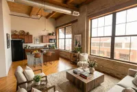 Chic loft-style living space with exposed brick walls, featuring a cozy seating area, modern kitchen with island, and large industrial windows providing ample natural light.