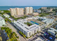 Aerial view of a vibrant seaside apartment complex with a modern design, featuring a central courtyard, lush greenery, and rooftop tennis courts, offering stunning ocean views.