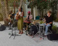 Vibrant outdoor band performance with a singer-guitarist, keyboardist, bassist, and drummer under lush palm trees.