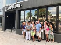 A group of cheerful people posing in front of a modern clam bar with large windows, reflecting a sunny day with palm trees in the background.