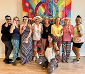 Joyful group of people embracing a retro style, wearing colorful and patterned outfits with peace signs and flower headbands, posing in front of a vibrant peace symbol backdrop.