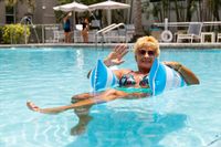 Elegant poolside relaxation with a smiling woman enjoying a sunny day on a float in a crystal-clear swimming pool, surrounded by palm trees and lounge chairs.