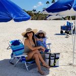 Two women smiling and relaxing on a sunny beach under blue umbrellas, enjoying a perfect day with sandy shores and clear skies.