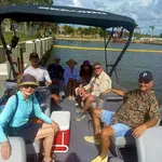 Group of people enjoying a sunny boat ride with smiles and casual attire, set against a serene dockside backdrop.