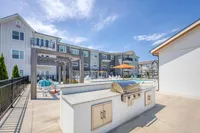 Modern apartment complex with outdoor grilling area, poolside seating, and vibrant orange umbrellas under a clear blue sky.