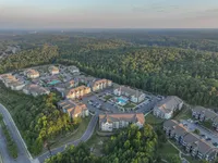 Aerial view of a modern apartment complex surrounded by lush greenery, featuring multiple buildings, ample parking, and a central swimming pool.