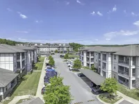 Sunny aerial view of a modern apartment complex with ample parking, lush greenery, and scenic surroundings.