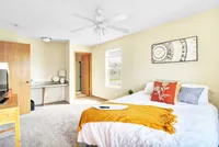 Bright and cozy bedroom with stylish decor and natural light, featuring a plush bed with colorful pillows, work desk, and soft carpet flooring.