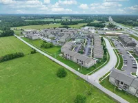Charming aerial view of a modern apartment community, surrounded by lush greenery and spacious fields, showcasing convenient road access and ample parking.