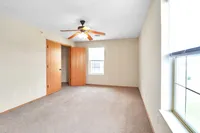 Bright and spacious bedroom with soft carpet flooring, featuring a ceiling fan, large window, and an open closet with wooden doors.